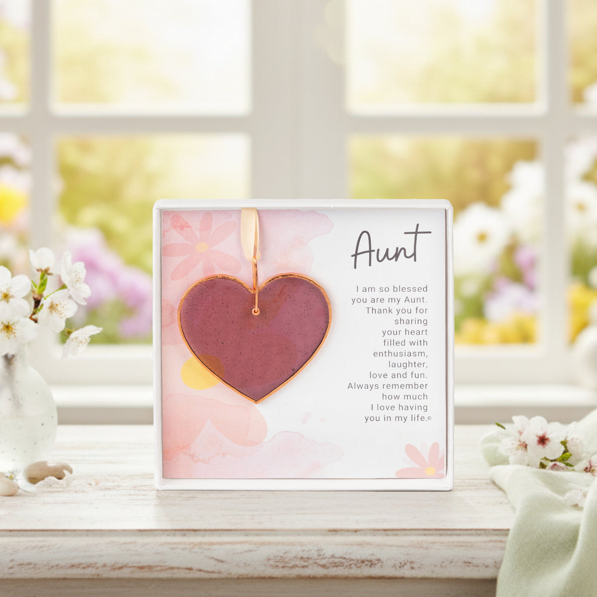 Stained glass rose mauve heart ornament packaged with "Aunt" sentiment in a white box with a clear lid sitting on a table in front of a window.