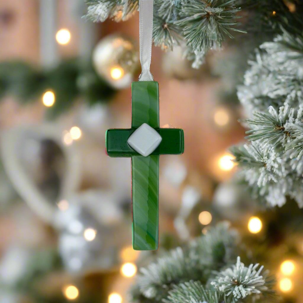 Green glass cross with white satin ribbon hanging on a Christmas tree.
