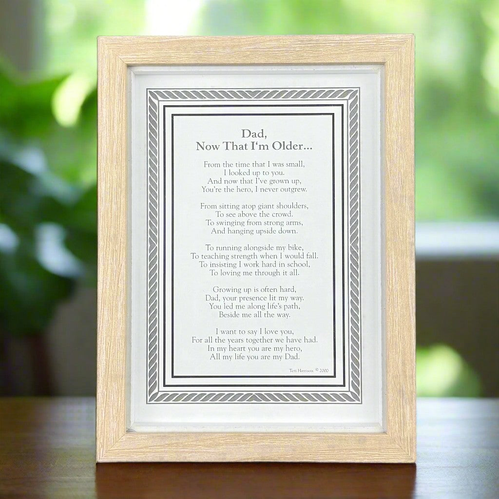 5x7 framed poem titled 'Dad, Now That I'm Older' on a wooden table in front of a garden window.