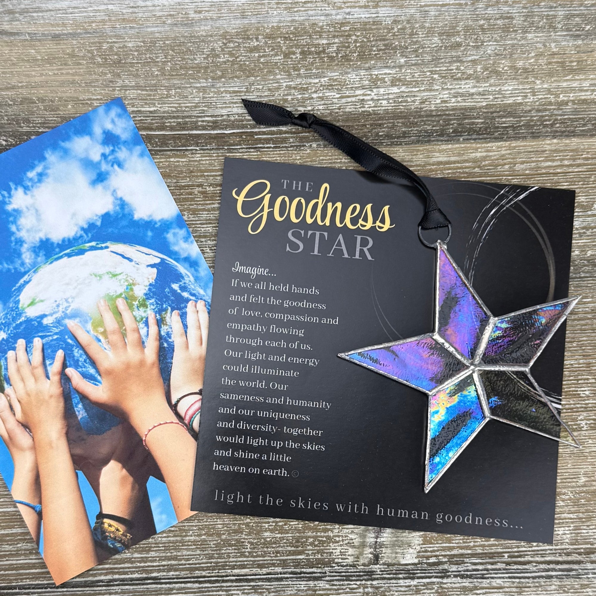 Handmade 4" clear iridescent stained-glass star with silver edging, packaged with "The Goodness Star" sentiment on a table with many hands holding a globe.
