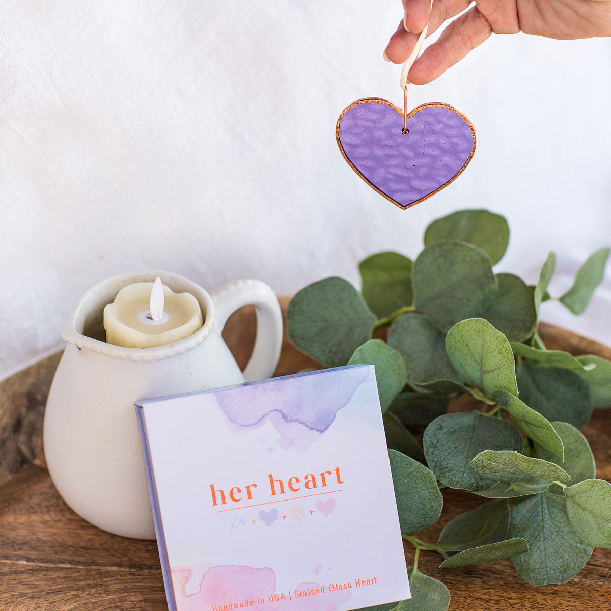 Purple stained-glass heart being suspended by a hand allowing the light to shine through and show the wavy texture and the color of the glass.